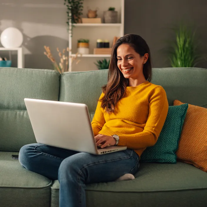 Eine lächelnde Frau mit langen braunen Haaren sitzt entspannt auf einem mintgrünen Sofa und arbeitet an einem Laptop.