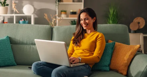 Eine lächelnde Frau mit langen braunen Haaren sitzt entspannt auf einem mintgrünen Sofa und arbeitet an einem Laptop.