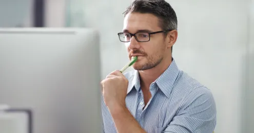 Mann mit Brille und Dreitagebart sitzt nachdenklich vor einem Computerbildschirm und hält einen Stift ans Kinn.