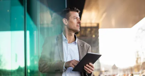 Mann im Businessanzug hält ein Tablet und blickt nachdenklich in die Ferne, stehend vor einem modernen Glasgebäude.
