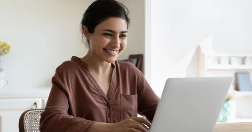 Lächelnde junge Frau sitzt an einem Holztisch und arbeitet an einem Laptop in einem hellen Raum
