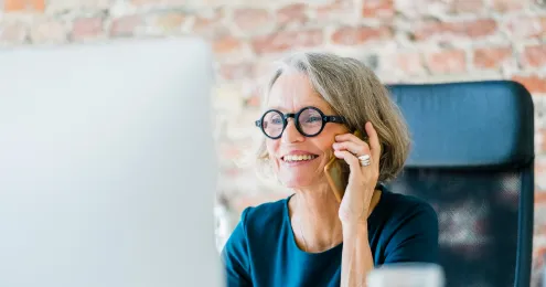 Lächelnde ältere Frau mit Brille telefoniert und sitzt vor einem Computer in einem modernen Büro mit Backsteinwand im Hintergrund.