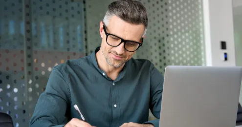 Mann mittleren Alters mit grau-braunem Haar und schwarzer Brille schreibt konzentriert etwas auf ein Blatt Papier, neben ihm ein aufgeklappter Laptop auf dem Schreibtisch.