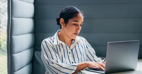 Junge Frau mit hochgestecktem dunklem Haar und weißem Streifenhemd sitzt konzentriert an einem Laptop in einer modernen Büro-Lounge.