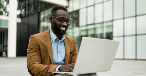 Lächelnder Mann mit Brille und braunem Sakko arbeitet mit einem Laptop im Freien vor einem modernen Bürogebäude