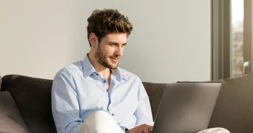 Junger Mann mit lockigem Haar und hellblauem Hemd sitzt lächelnd auf einem Sofa und arbeitet an einem Laptop