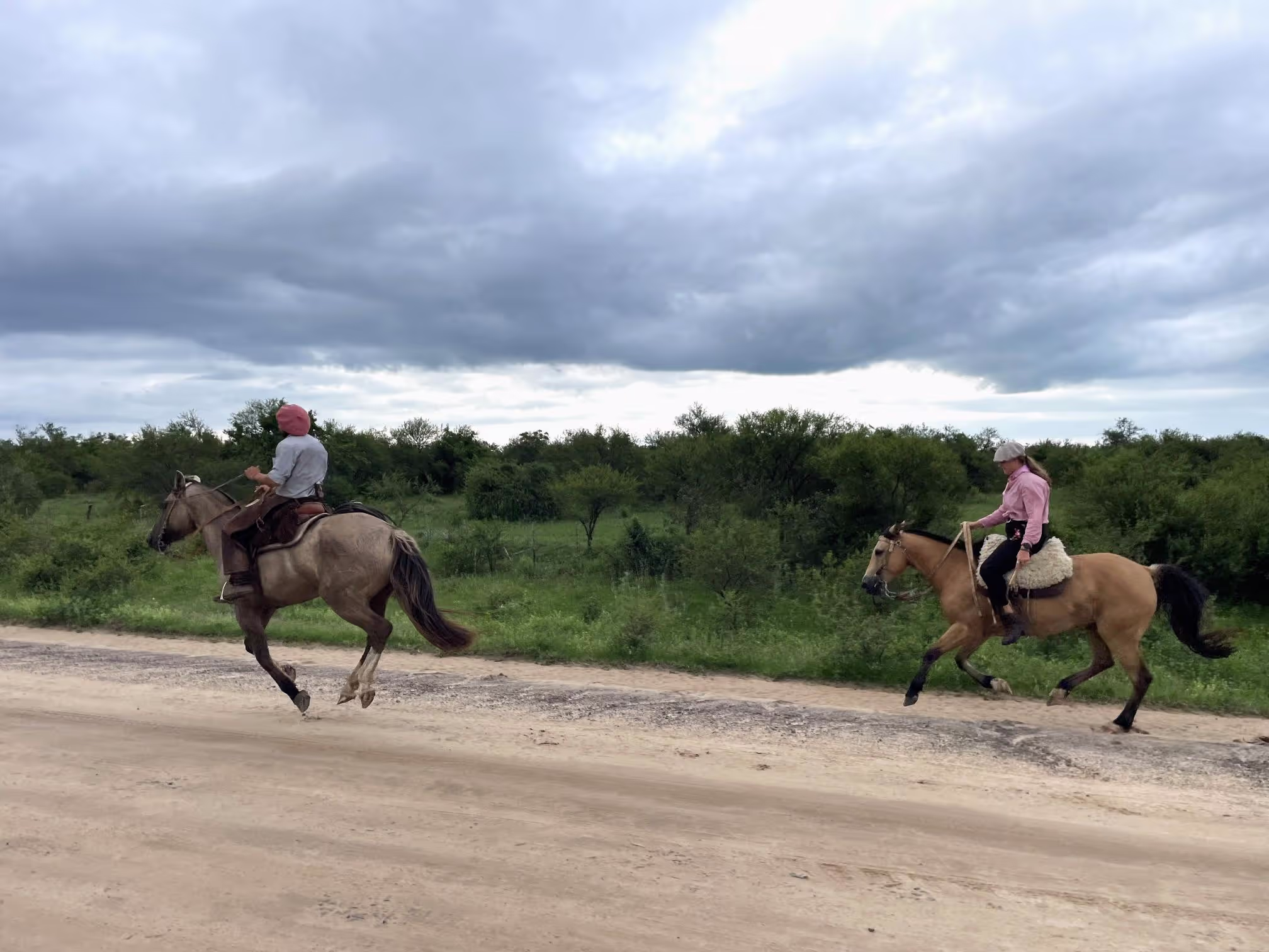 Two horses and gauchos galloping on a dirt road with green bushes and fields in the background