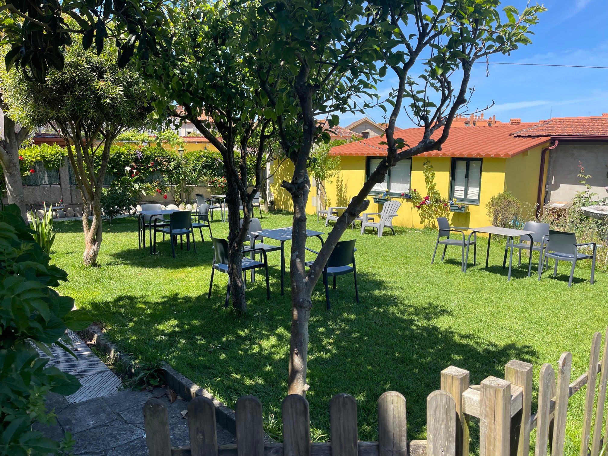 Yellow house with green garden full of trees and tables with chairs to relax in the sun