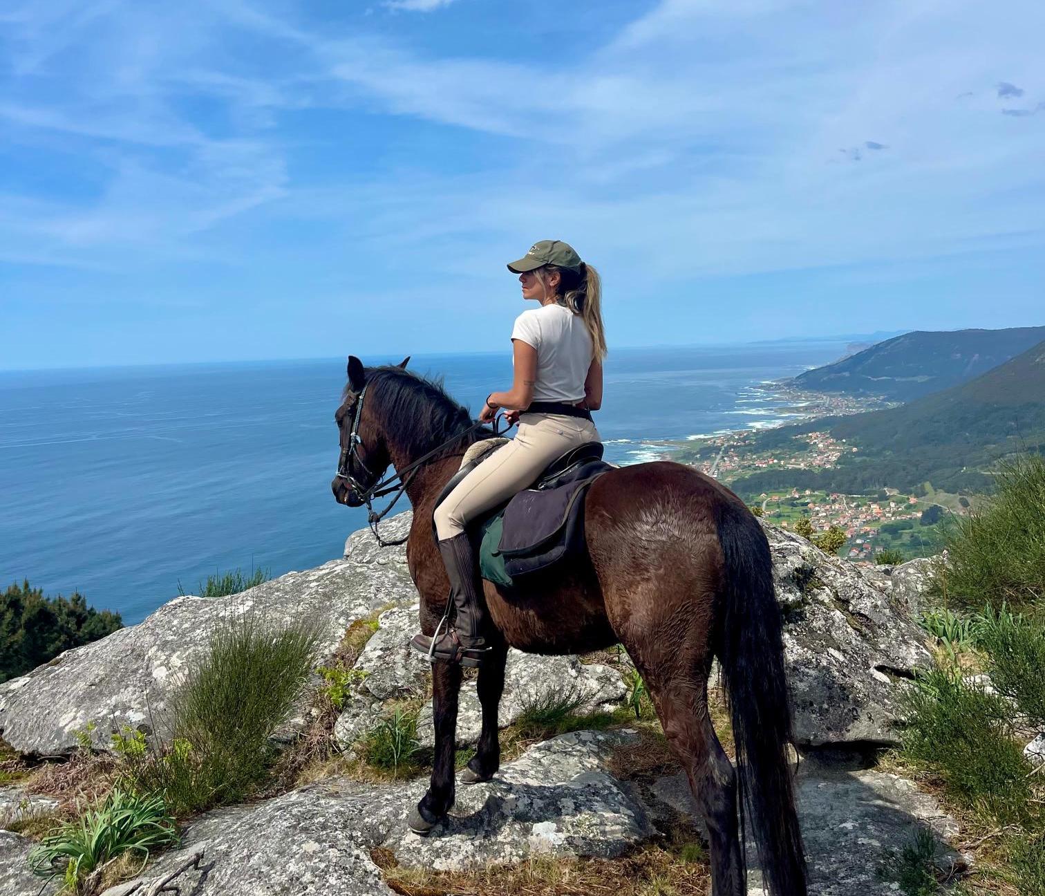 Woman on black horse, standing on the edge of the mountains, overlooking the ocean on a sunny day 