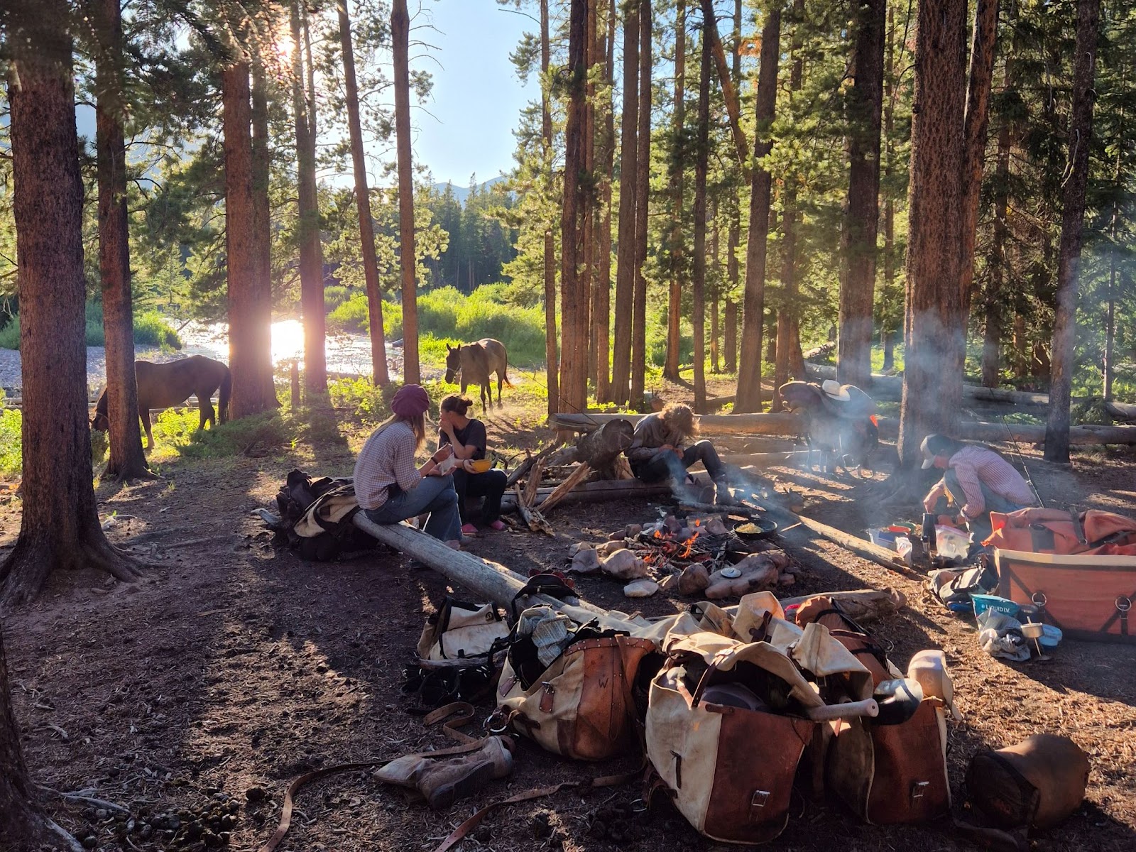 Group of riders sitting around a campfire while horses graze around them at sunset in the forest with a river running nearby
