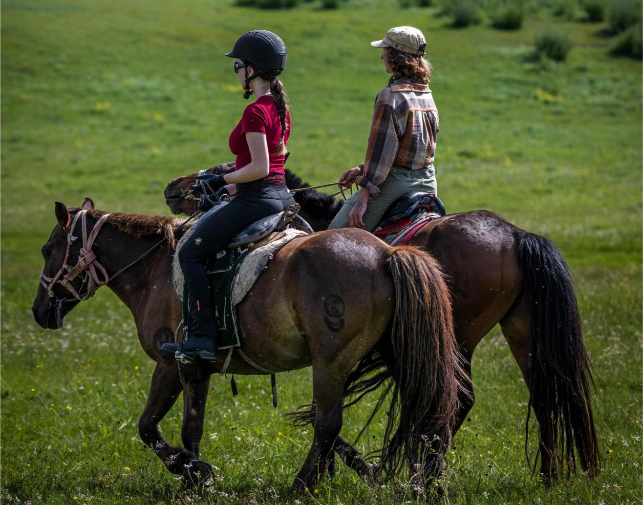 Two women riding semi-wild mongolian horses in a green grass field