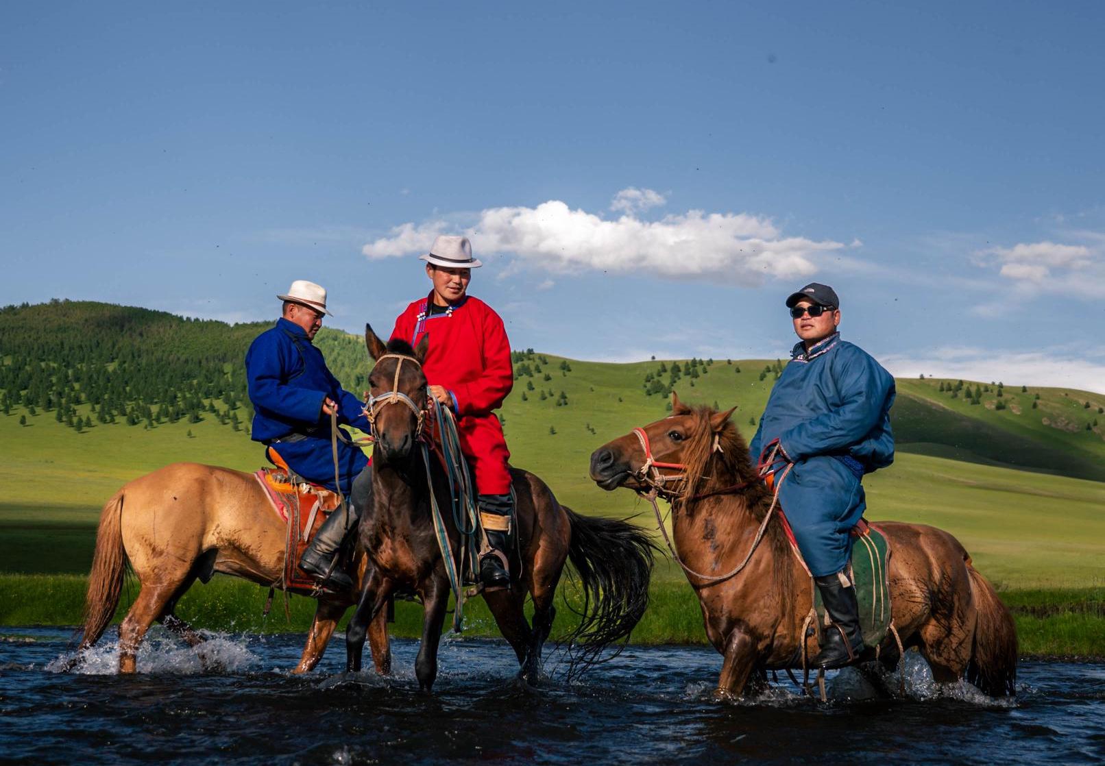 Mongolian men riding their horses through a river surrounded by green grass and hills on a sunny day