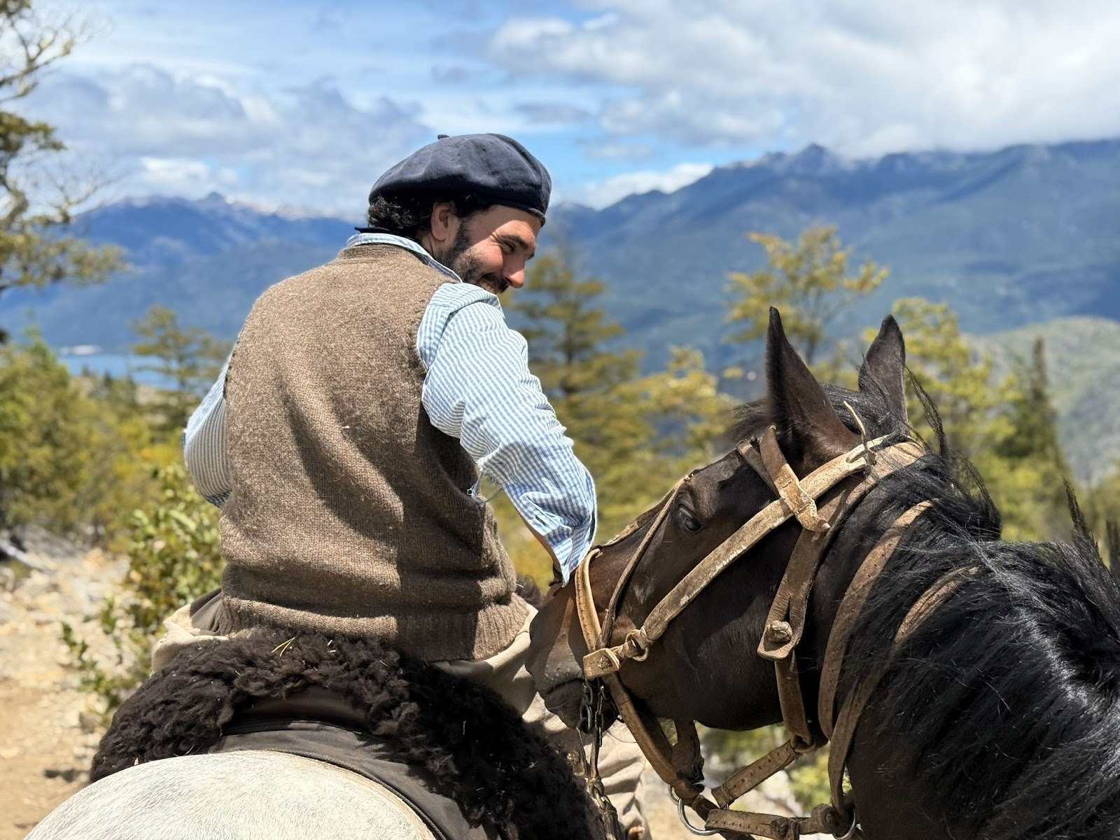 Gaucho on horseback smiling at black horse behind him on a narrow mountain trail next to a river