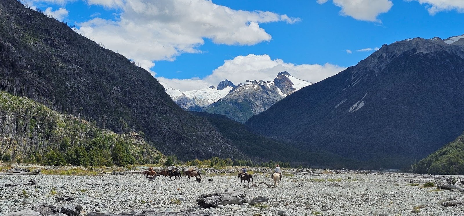 Riders on horseback on open rocky field in front of snow capped mountains