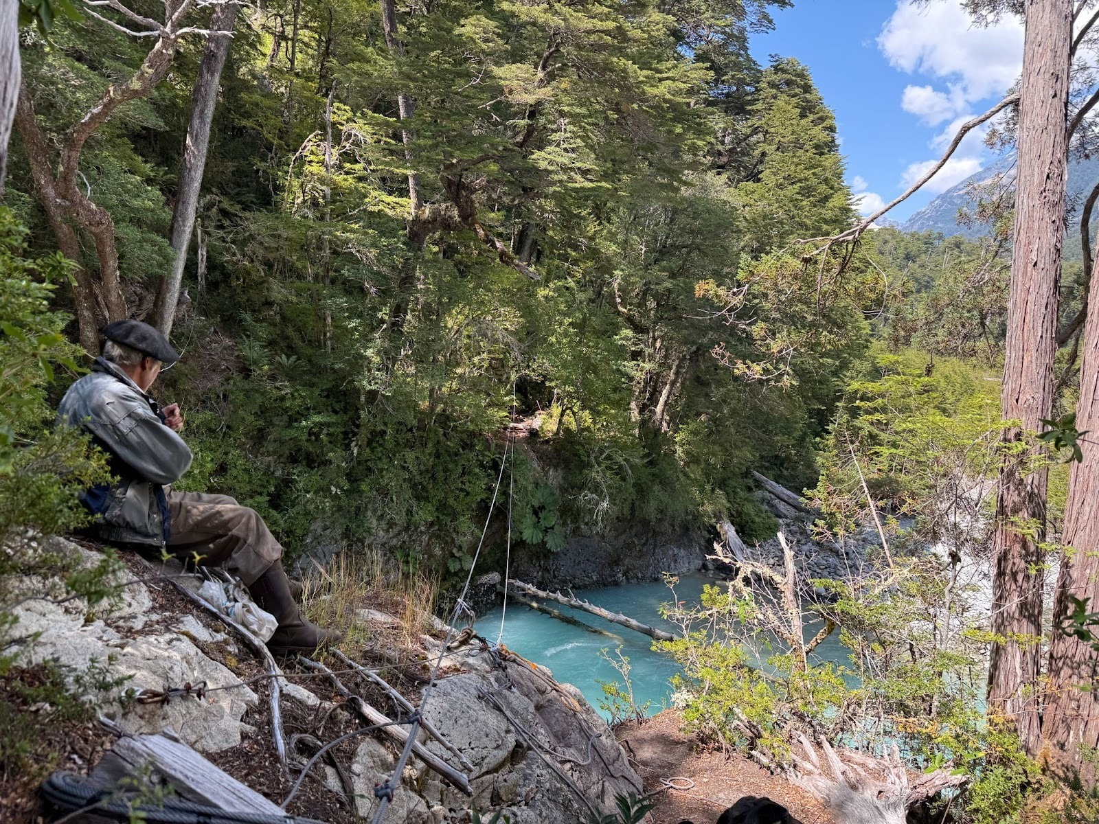 Gaucho sitting on a rock overlooking the forest and river, with mountains in the background on a sunny day