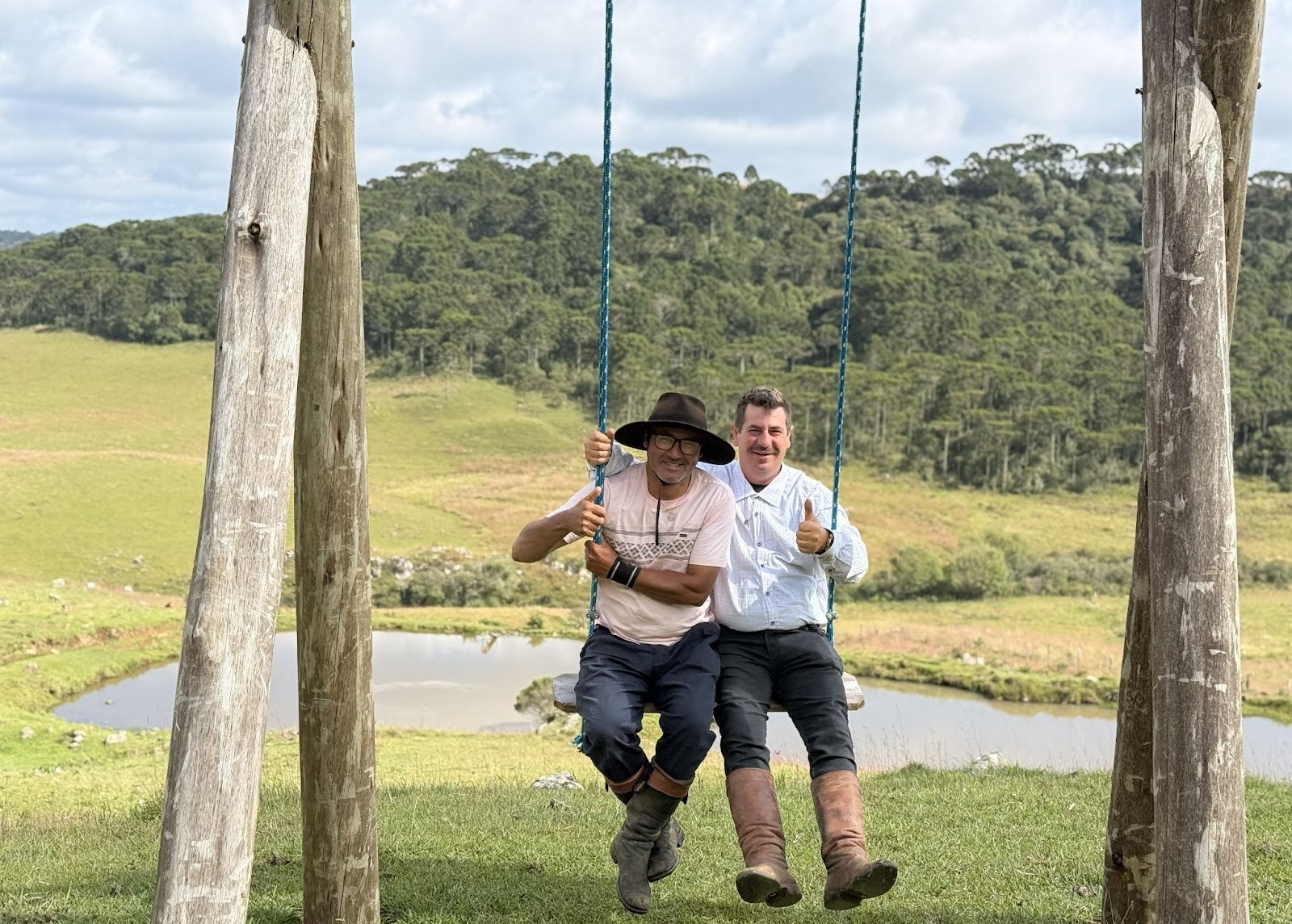 Two men on a swing in front of a pond surrounded by green fields and forest in the background 