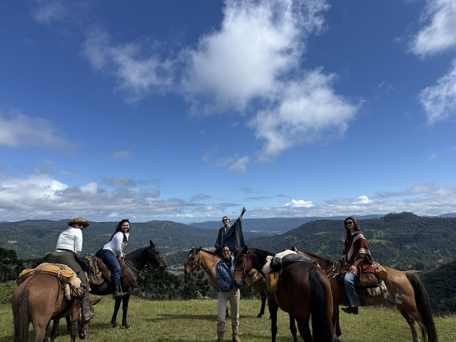 Group of riders on horseback posing for a picture in a green field in front of a view of the valley and mountains below