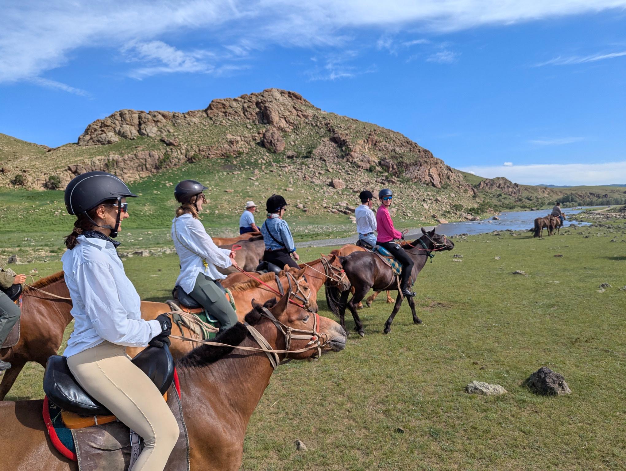 Group of riders on Mongolian horses walking along a river surrounded by hills on a sunny day