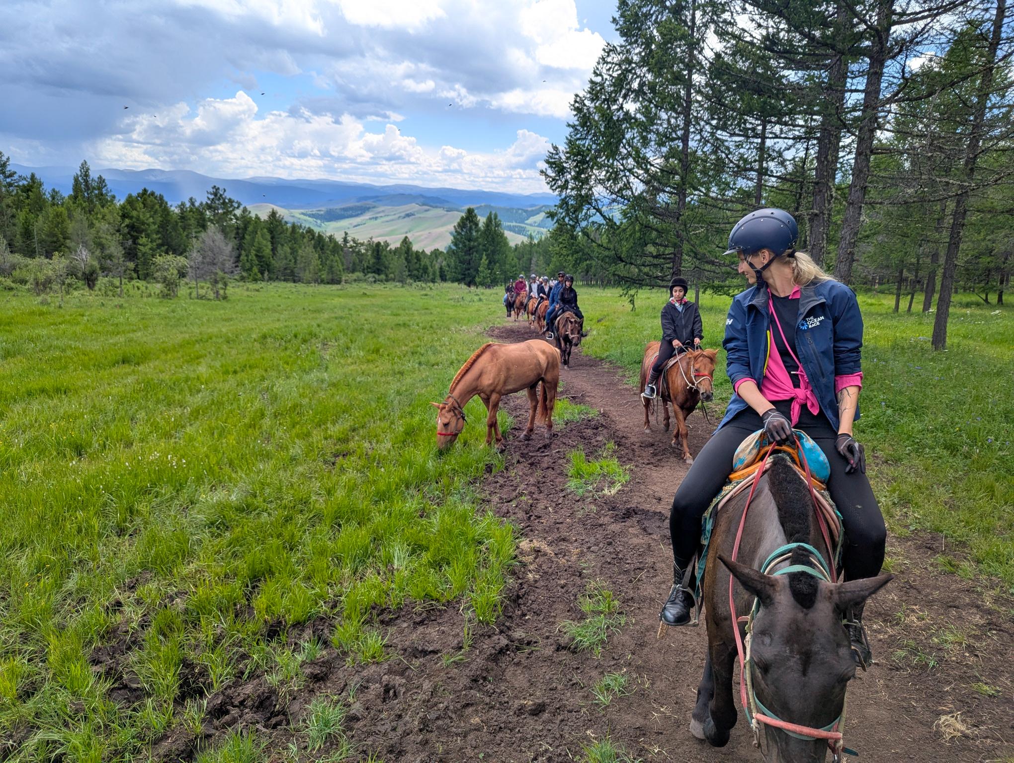 Group of riders on horses walking in a line along a dirt road in the forest, with green hills in the background