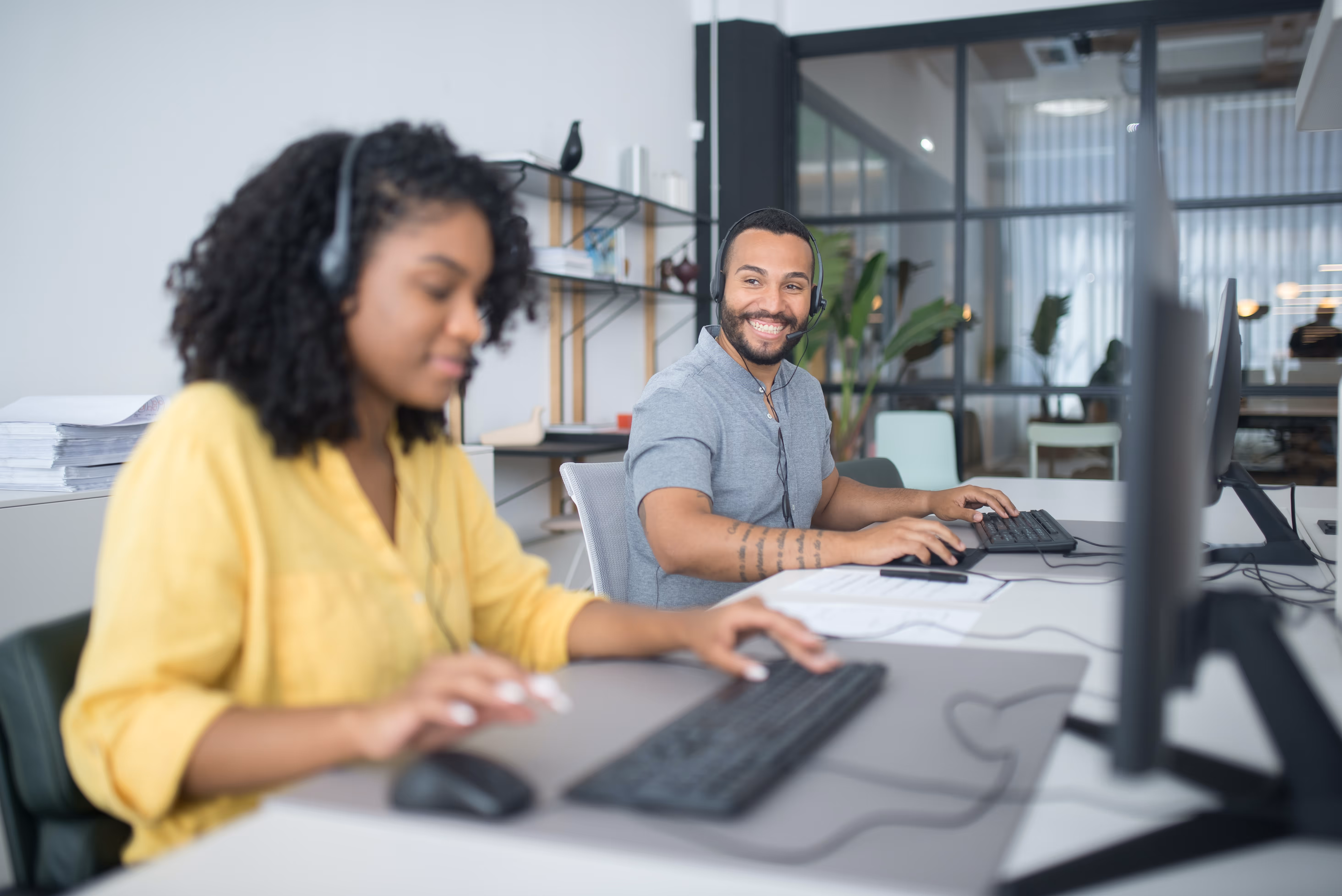 Deux professionnels travaillant sur des ordinateurs dans un bureau moderne et lumineux, l'homme à droite souriant à la caméra avec un casque tandis que la femme à gauche se concentre sur son écran, illustrant une atmosphère de travail collaborative et efficace.