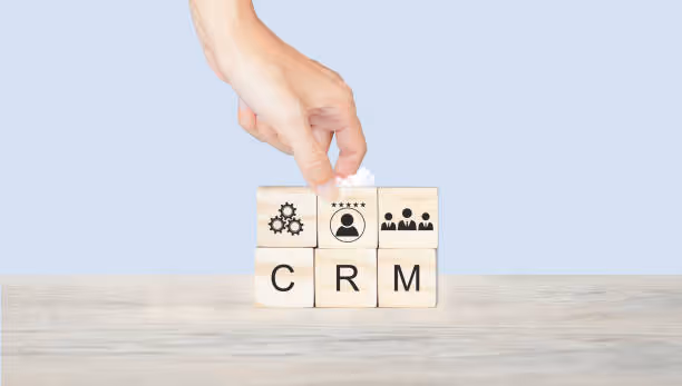 Une main humaine place la dernière pièce d'un puzzle sur des blocs en bois illustrant les lettres "C R M" pour Customer Relationship Management, avec des icônes représentant l'engrenage de la technologie, un service client de haute qualité, et une communauté d'utilisateurs, évoquant une stratégie de gestion de la relation client efficace et intégrée sur un fond bleu neutre.