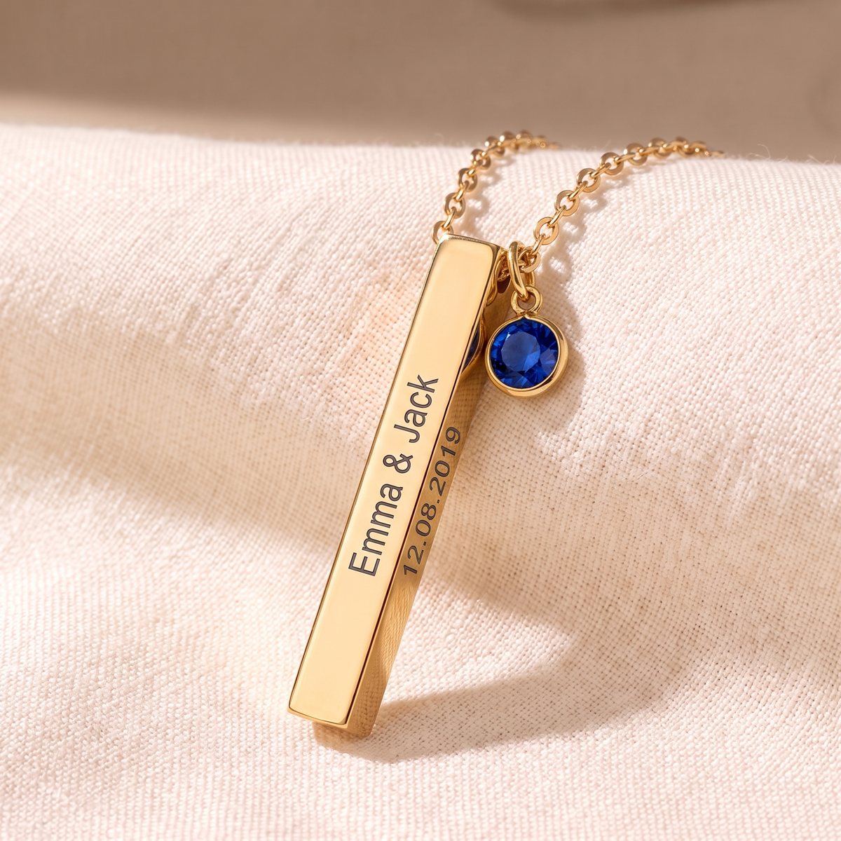 Gold 3D bar necklace engraved with Emma and Jack and a date, with a blue birthstone charm, on linen