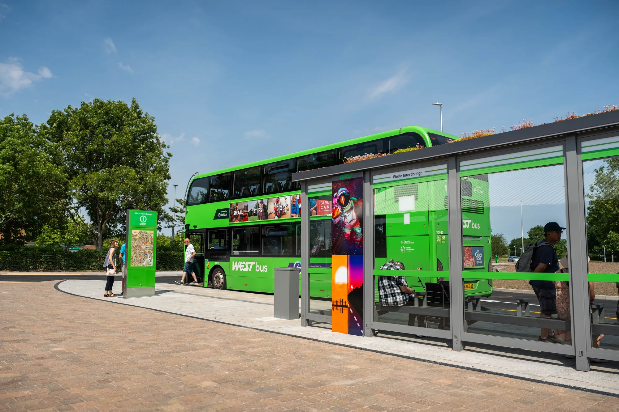 A green WEST double-decker bus parked beside a glass shelter as passengers board, viewed from the paved sidewalk.