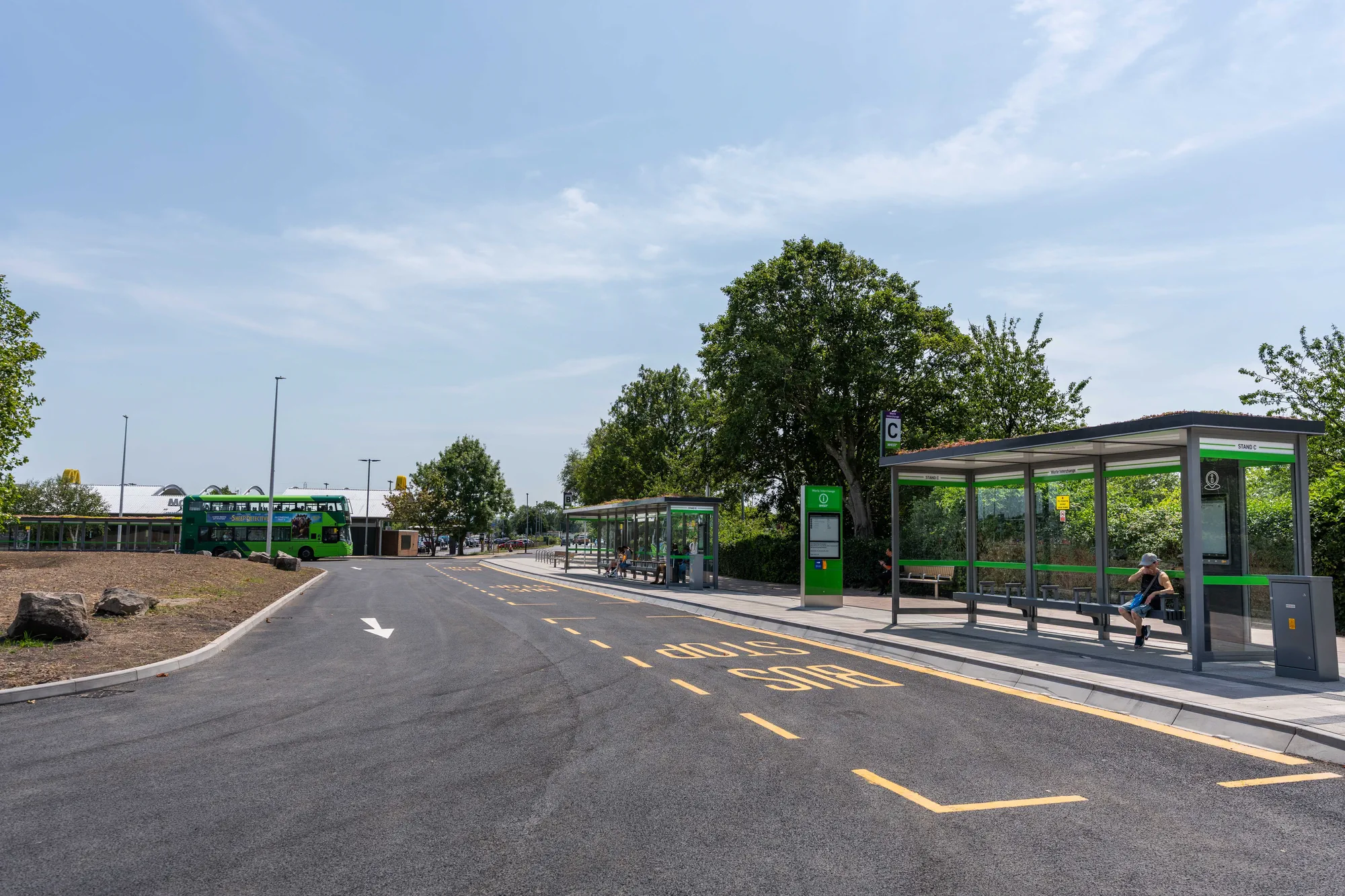 Wide view of the Worle Interchange bus bay showing paved lanes with yellow road markings, glass shelters, and a green bus in the distance.