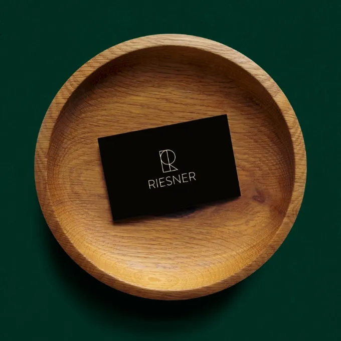 Black business card with white logo and text 'RIESNER' placed inside a round wooden bowl on a dark green background.