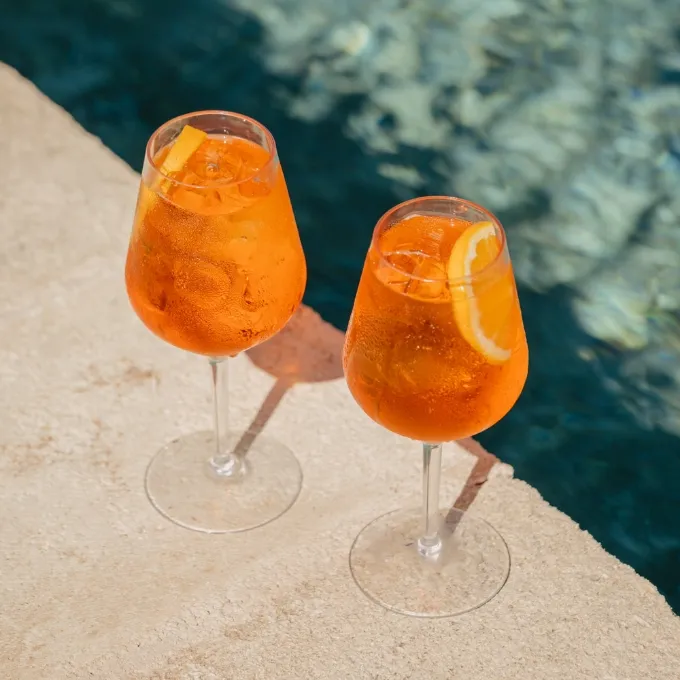 Two orange-colored cocktails with ice and orange slices on a poolside ledge with water in the background.