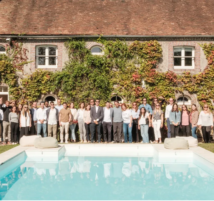 Group of people standing in a line beside a swimming pool with a brick building covered in ivy in the background.