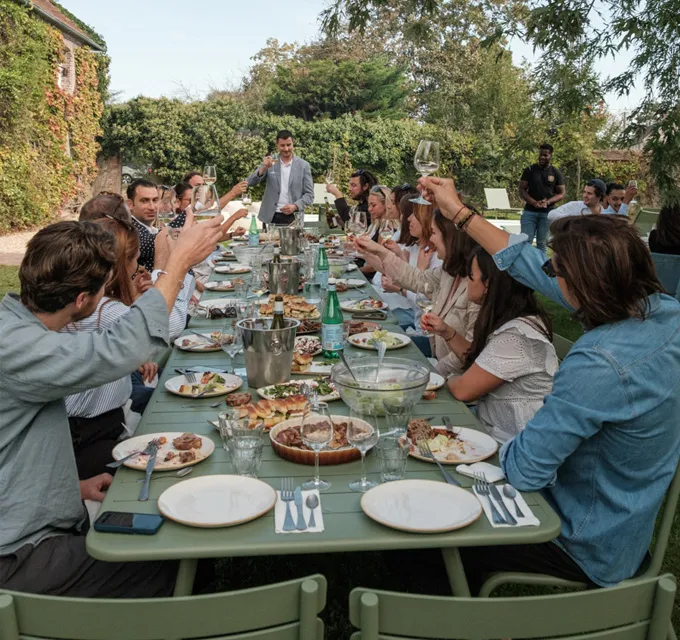 A group of people seated around a long outdoor table raising wine glasses for a toast during a garden gathering.
