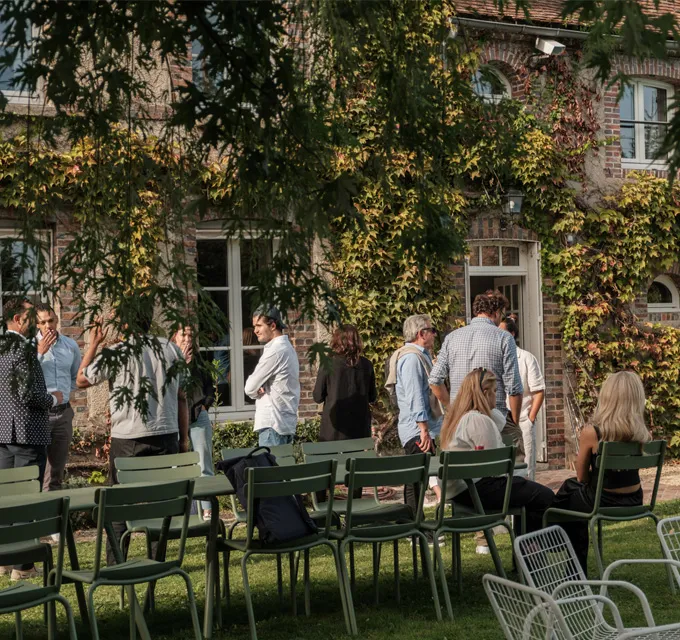 People socializing outdoors near a brick building covered with green ivy, with empty green chairs in the foreground.