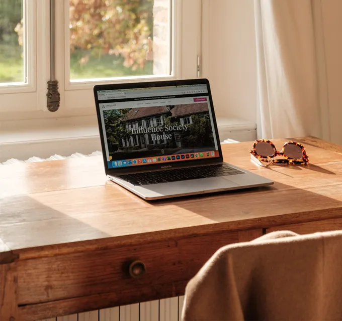 A laptop on a wooden desk near a window with sunglasses beside it.