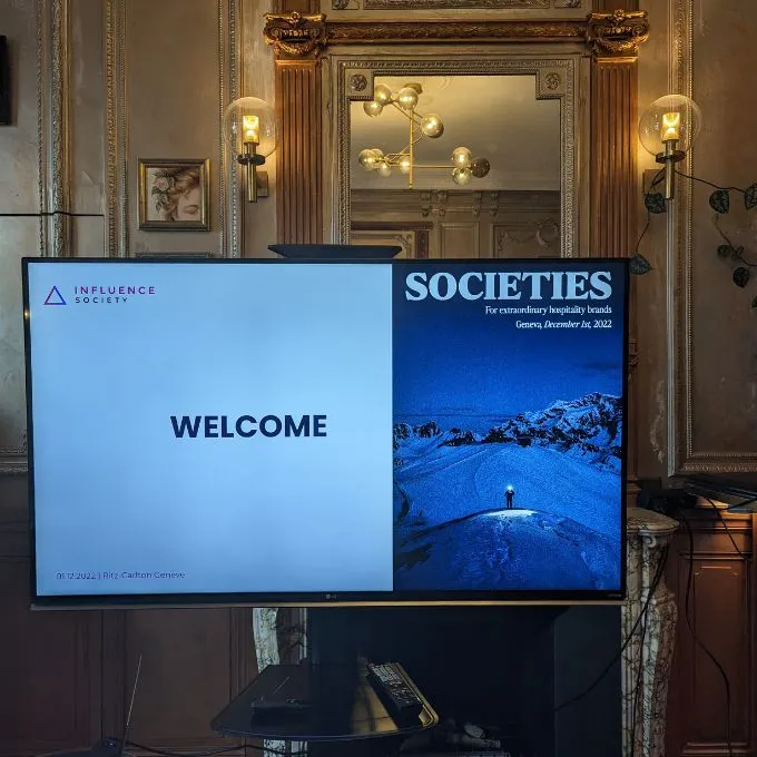 Large screen in an ornate room displaying a welcome slide for Influence Society's Societies event in Geneva on December 1st, 2022, with a snowy mountain image.
