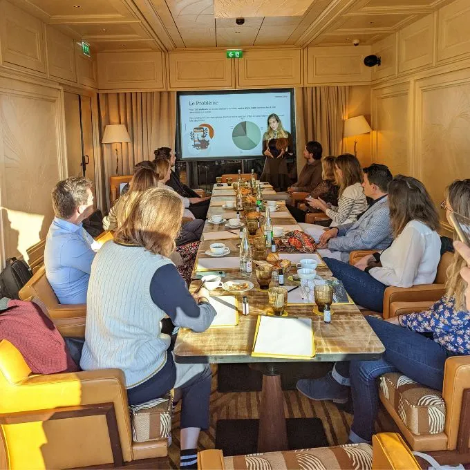Group of people seated around a conference table in a wood-paneled room, attentively listening to a woman presenting data with pie charts on a screen.