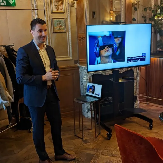 Man in a dark suit giving a presentation in a room with classic decor, with a large screen showing a person wearing a virtual reality headset.