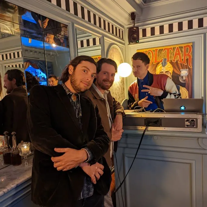 Three men posing together indoors near a DJ booth with mirrors and warm lighting.