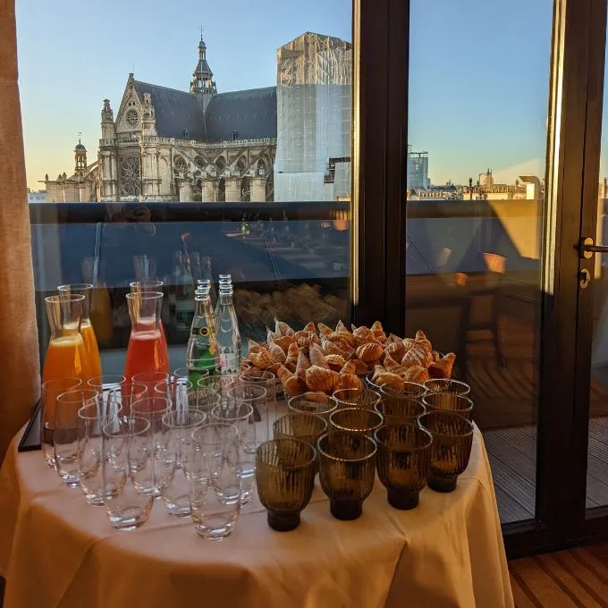 Table with glasses, bottled drinks, juice, and croissants set in front of a large window showing a Parisian church and cityscape at sunset.