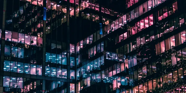 Glass facade of a modern office building at night with illuminated interiors reflecting various colors.