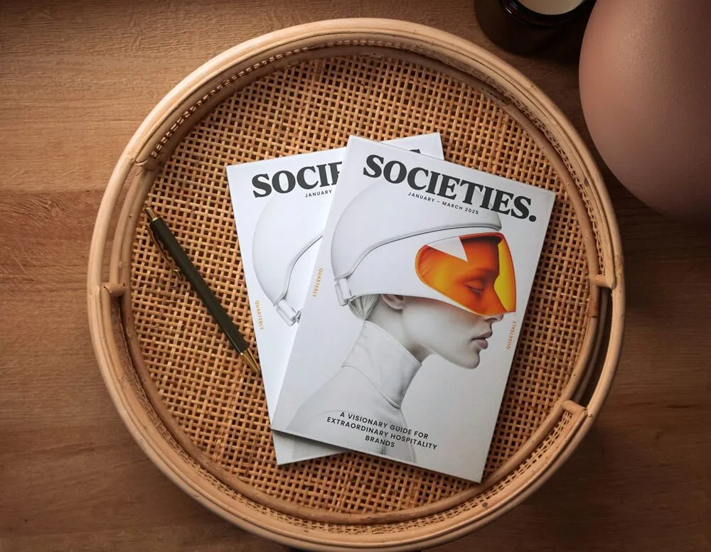 Two copies of SOCIETIES magazine with a futuristic woman on the cover, placed on a woven rattan tray with a pen.