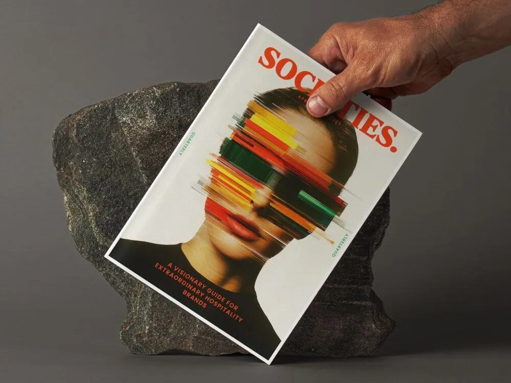 A hand holding a magazine titled 'SOCIETIES' featuring a woman's face with colorful glitch-like horizontal stripes over her eyes.