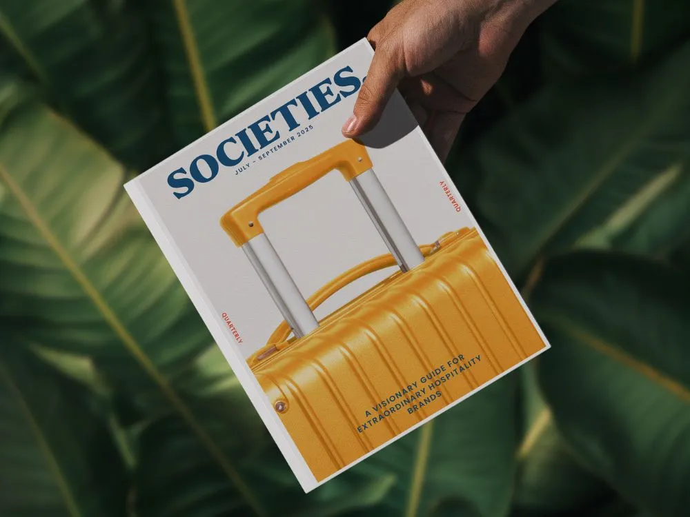 Hand holding a magazine titled 'SOCIETIES' featuring a yellow suitcase handle on the cover.