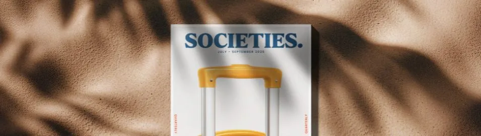 Top view of a magazine titled 'SOCIETIES.' on sandy beach with shadows of palm leaves.