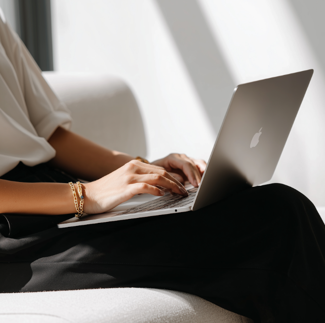 Person typing on a silver Apple laptop while sitting on a light-colored sofa in bright natural light.