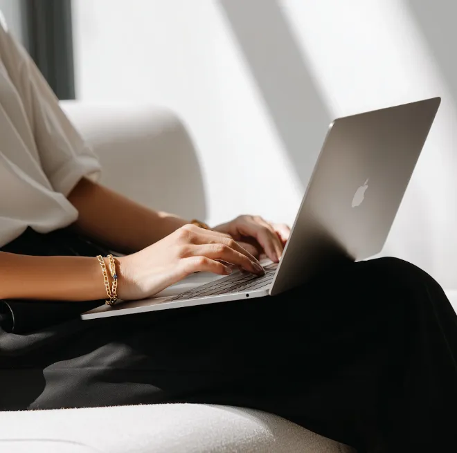 Person typing on a silver Apple laptop while sitting on a light-colored sofa in bright natural light.