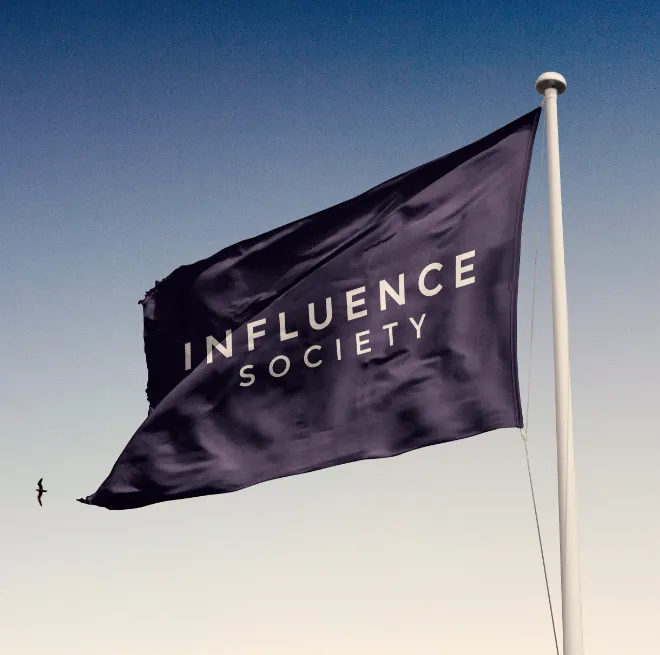 Dark flag with the words 'INFLUENCE SOCIETY' waving on a tall white flagpole against a clear sky background with a bird flying nearby.
