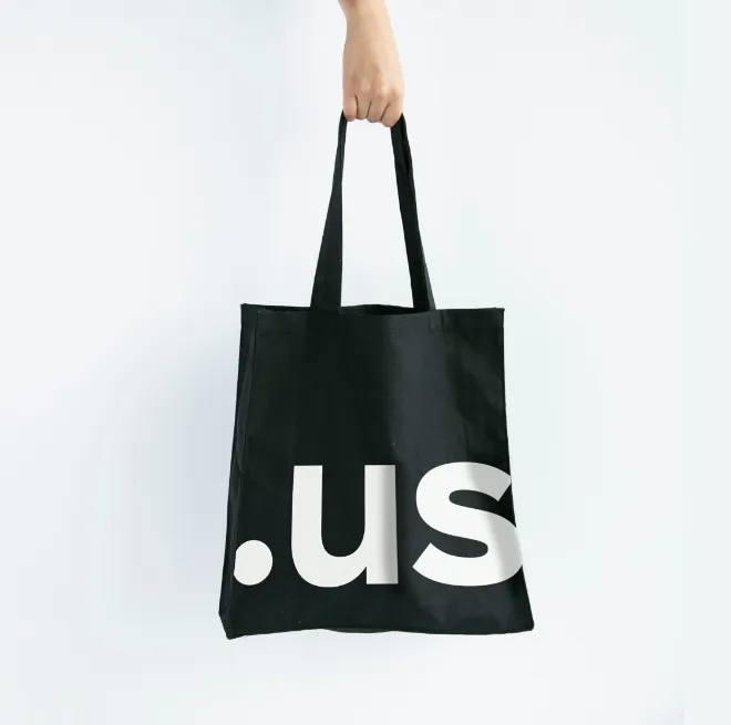 Hand holding a black tote bag with large white text reading '.us' on it.
