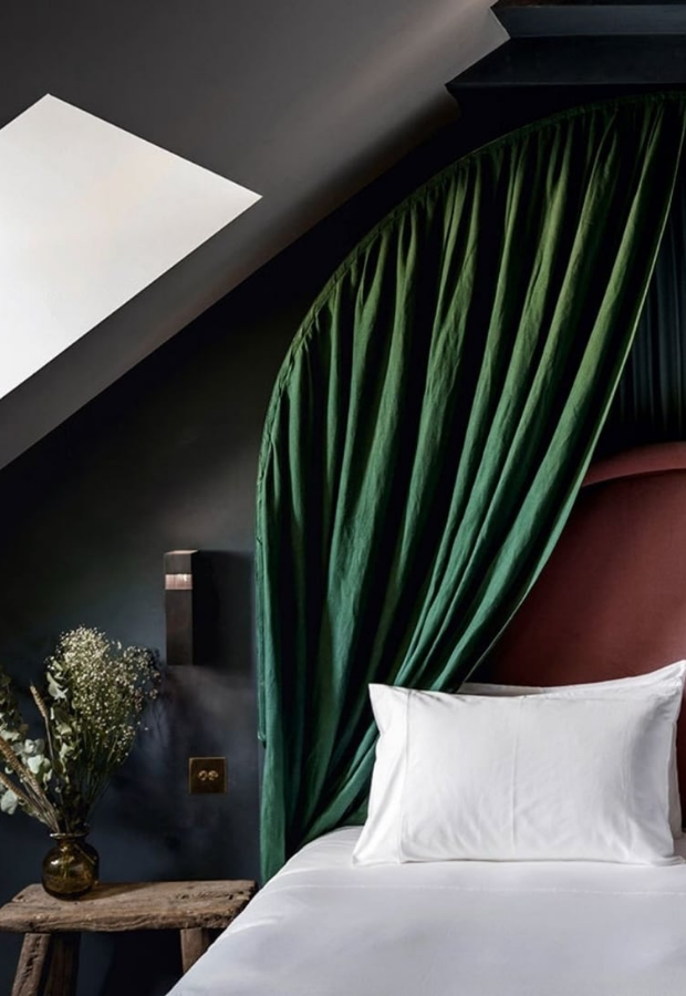 Cozy bedroom corner with white pillow and bed linens, green draped curtain canopy, dark walls, and a rustic wooden stool with a vase of dried flowers.