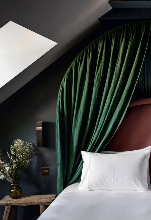 Cozy bedroom corner with white pillow and bed linens, green draped curtain canopy, dark walls, and a rustic wooden stool with a vase of dried flowers.